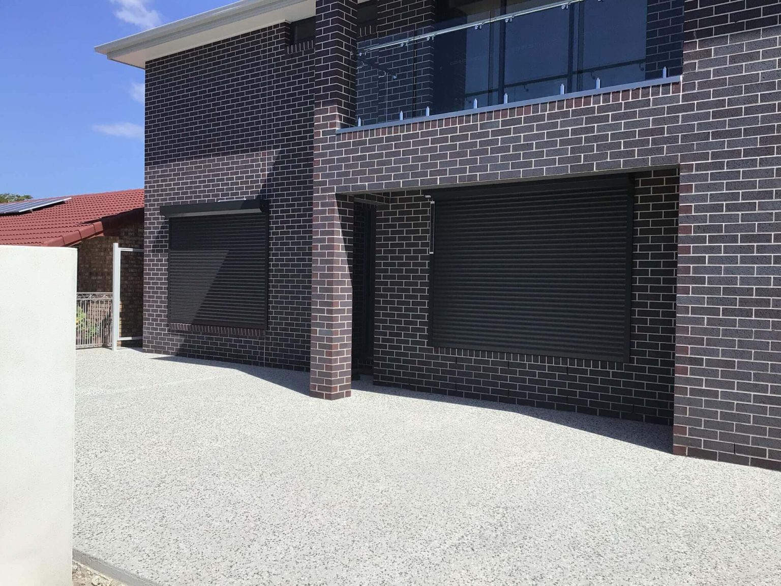 A cream roller shutter fully closed over a wide window on a cream brick single-storey home, with surrounding garden beds and a lawn in the foreground.