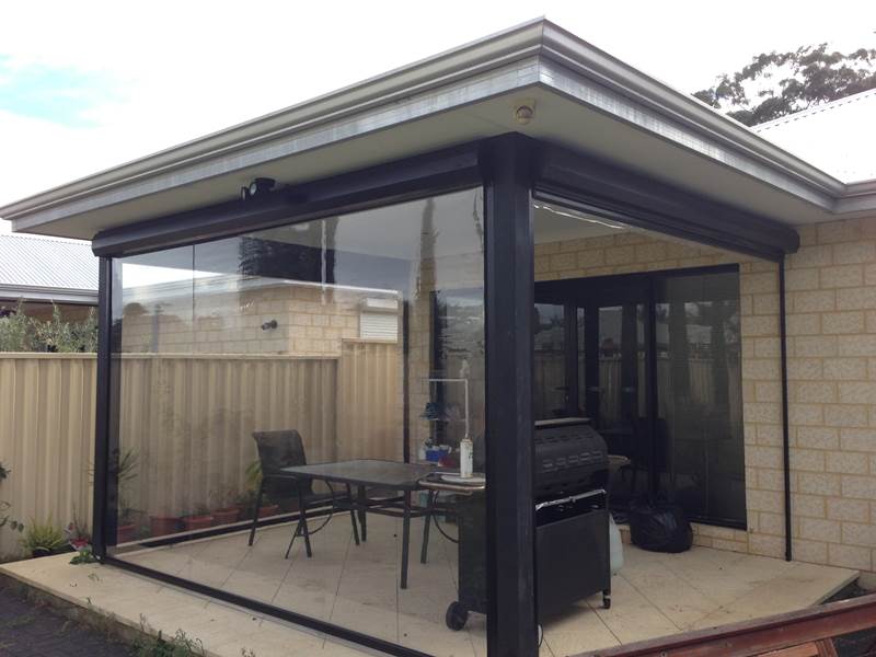 Clear PVC café blinds fully enclosing a freestanding pergola alfresco with outdoor dining setting and BBQ on limestone paving, attached to a cream limestone brick home.