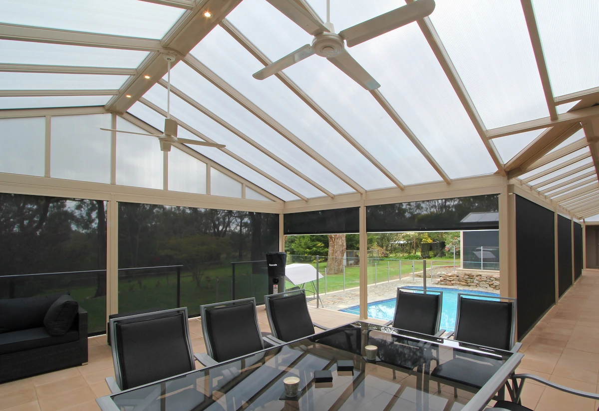 Dark charcoal slide-track outdoor blinds fully lowered around a large gabled glass-roof alfresco enclosure, with black outdoor dining furniture, a pool, and garden beyond, photographed from inside.