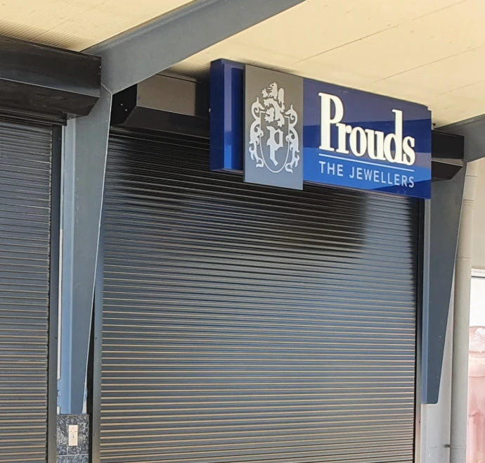 A close-up of a charcoal solid defender security roller shutter closed over the entrance of Prouds The Jewellers, with the branded blue and grey signage prominently displayed above.