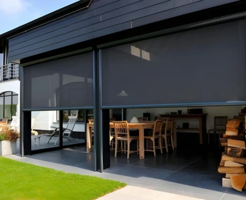 Three charcoal zip-track outdoor blinds partially lowered across a large glazed alfresco dining area on a dark weatherboard home, with timber dining furniture visible inside and a lawn beyond.